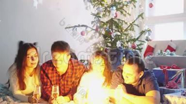 快乐的年轻朋友躺在圣诞树旁的地毯上在装饰好的房间里点燃火花庆祝新年